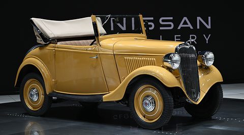 A yellow two-seat roadster with its white hood folded, a rounded grille and spare wheel, before a dark wall. Opens a larger view