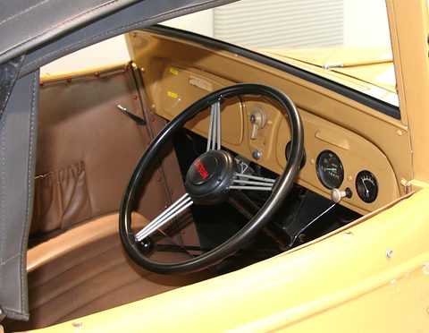 A black four-spoke steering wheel in front of a yellow painted dashboard with round dials. Opens a larger view