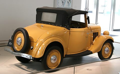 The rear of a yellow roadster with its black hood up and a spare wheel on the rounded tail. Opens a larger view