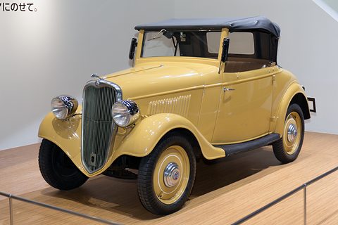 A yellow two-seat roadster with a dark blue hood up and a chrome grille on a wooden show floor.