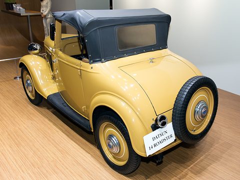 The rear of a yellow roadster with a dark blue hood, a spare wheel and a small plaque, on a wooden floor. Opens a larger view
