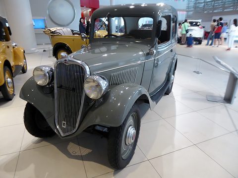 A small grey two-door saloon with a tall dark grille and round headlamps on a white gallery floor. Opens a larger view