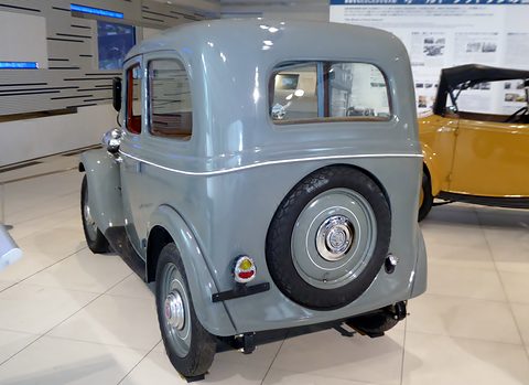 The rear of a small grey two-door saloon with a spare wheel on its back and a small rear window. Opens a larger view