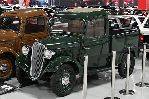 A small green pick-up truck with a chrome grille and black wings, a brown truck beside it in a garage hall. Opens a larger view