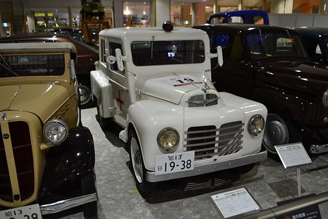 A small white two-door car with a horizontal-bar grille and round headlamps among other old cars in a museum. Opens a larger view
