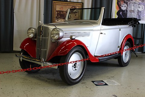 A silver open tourer with red wings and white-wall tyres behind a red rope in a showroom. Opens a larger view
