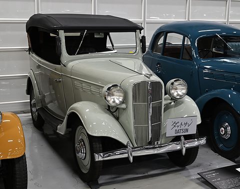A pale grey open tourer with its black hood up, a tall grille and white-wall tyres in a garage hall. Opens a larger view