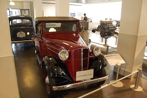 A red roadster with black wings and its tan hood up in a museum corridor, an engine display behind. Opens a larger view