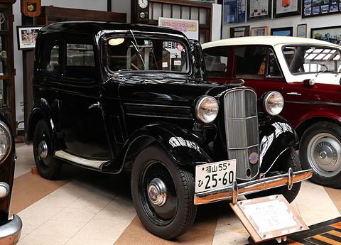 A small black four-window saloon with a chrome grille and a Japanese number plate in a museum room. Opens a larger view