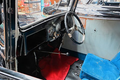 The cabin of an old saloon: a large steering wheel, round dials, red floor mat and blue upholstered seat. Opens a larger view
