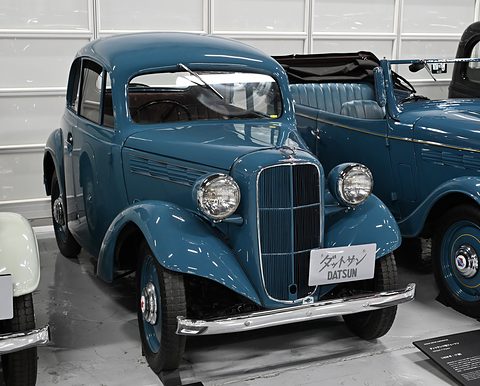 A blue two-door coupé with a chrome grille and round headlamps beside another blue car in a garage hall. Opens a larger view