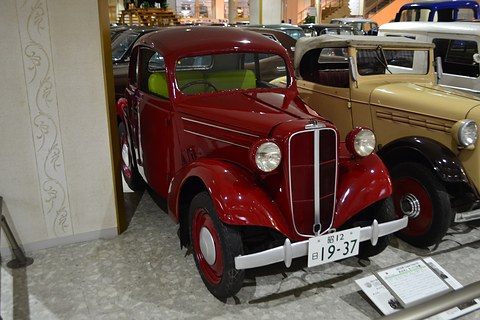 A red two-door car with black wings, a tall chrome grille and a Japanese number plate in a museum. Opens a larger view