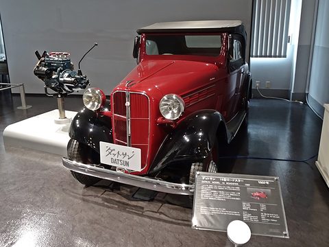 A red roadster with black wings and a beige hood beside a museum display card, an engine on a stand behind. Opens a larger view