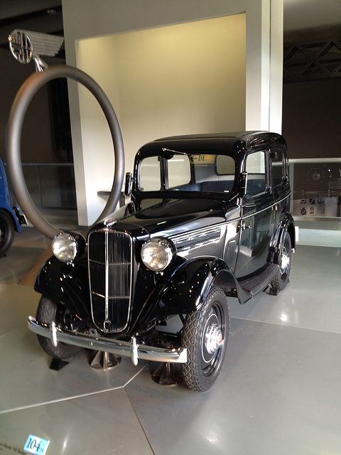 A small black four-window saloon on a pale museum floor beside a large ring-shaped sculpture. Opens a larger view