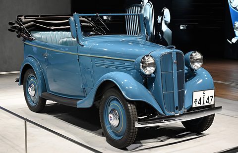 A blue open tourer with a tall grille, white-wall tyres and a folded black hood on a white plinth. Opens a larger view