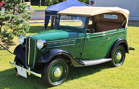 A dark green open tourer with a beige hood and black wings parked on a lawn by trees and a tent. Opens a larger view