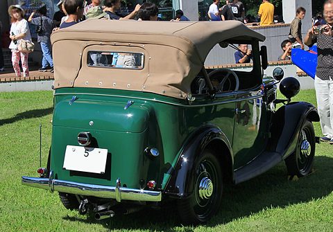 The rear of a green open tourer with a beige hood and a white number plate on grass, onlookers behind. Opens a larger view