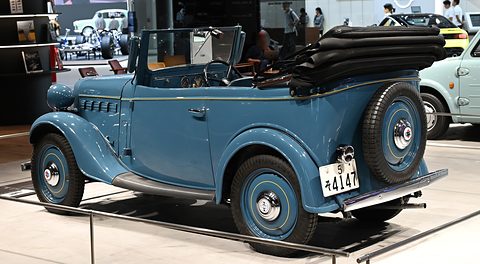 The rear of a blue open tourer with a folded black hood and spare wheel on a white plinth. Opens a larger view