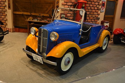 A blue roadster with bright yellow wings and wheels, its windscreen up and no hood, in a museum by a brick wall. Opens a larger view