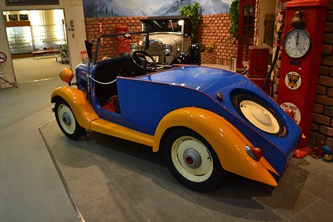 The rounded blue tail of a roadster with yellow wings and a covered spare wheel set into its back. Opens a larger view