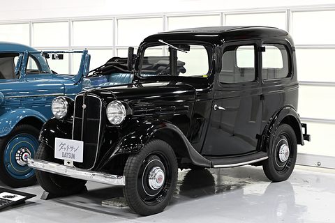 A small black four-window saloon with a chrome grille in a white garage hall. Opens a larger view