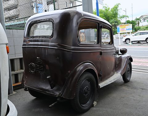 The rear of a small dark brown saloon with weathered paint and a spare-wheel mount, parked under cover by a street. Opens a larger view
