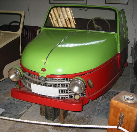 Head-on view of a green and red three-wheeler with a chrome grid grille, twin headlamps and a red bumper. Opens a larger view