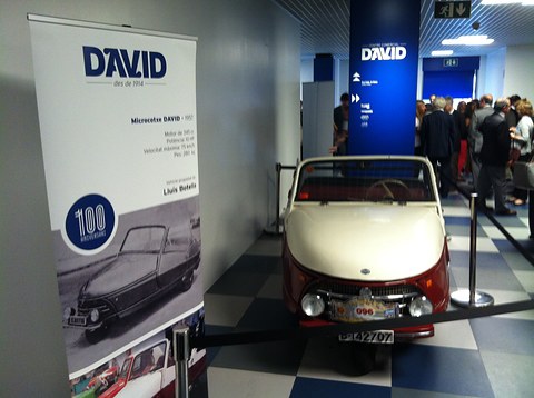 Cream and red three-wheeler behind a rope beside a banner reading DAVID with a 100 roundel, visitors in the hall.