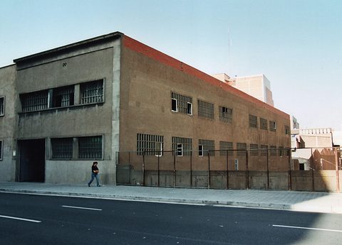 A plain two-storey brick factory with barred windows on a street corner, a woman walking past on the pavement. Opens a larger view