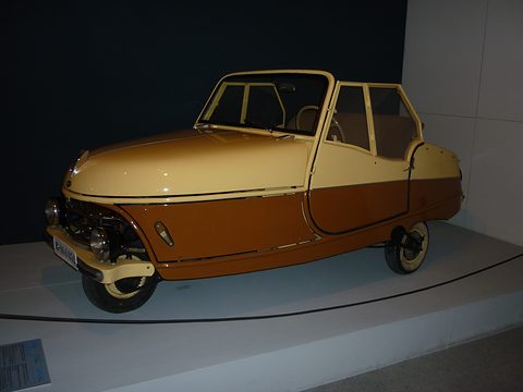 Cream and brown open three-wheeler with a rounded nose over a single front wheel, a windscreen and a folded hood. Opens a larger view