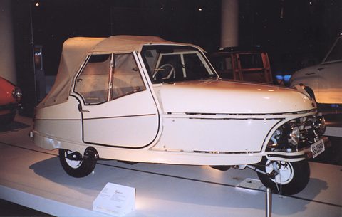 White three-wheeler with a beige hood and side screens on a museum plinth, lit against a dark background.