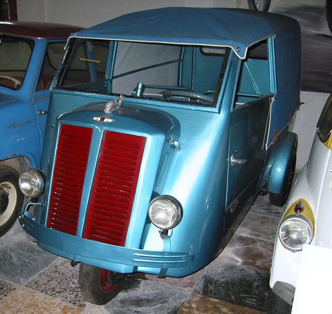 Front of a pale blue three-wheeled van with a red split grille and a canvas roof, among other old vehicles. Opens a larger view
