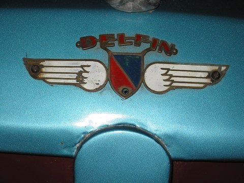 Winged enamel badge with a red and blue shield and the word DELFIN above, on pale blue metallic paint. Opens a larger view