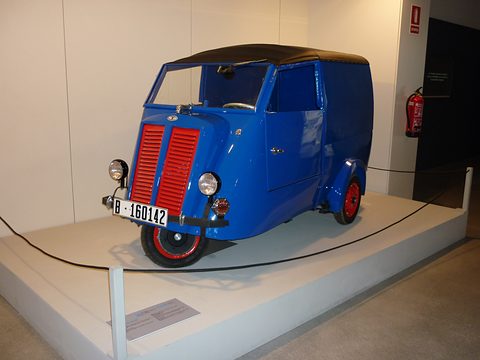 Blue three-wheeled van with a single red-hubbed front wheel and red louvred grille on a white plinth.