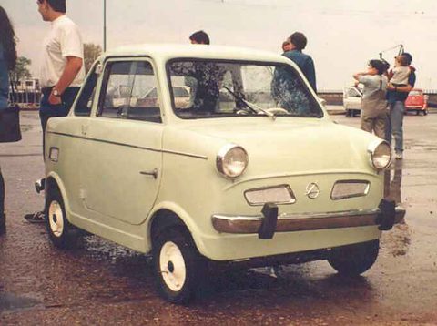 A small rounded pale green two-door car with air intakes in its nose and bumper over-riders, men standing round it.