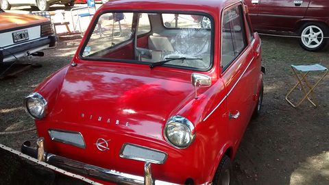 Red rounded small coupé with round headlamps, twin chrome air slots in its nose and a chrome bumper, parked on dirt beside folding chairs.