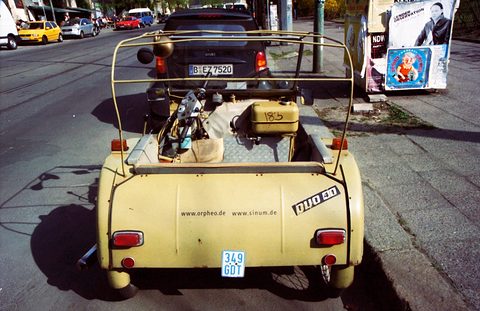 Rear of a cream three-wheeler with a bare hood frame and a DUO 4/1 badge, a black Smart car parked in front of it. Opens a larger view