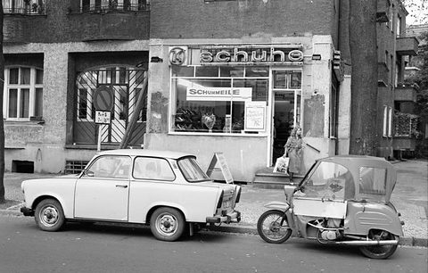 Black-and-white street scene: a hooded three-wheeler parked nose to tail with a Trabant before a shop front reading Schuhe.