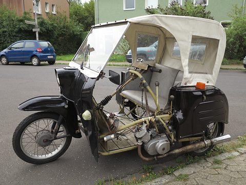 Black three-wheeler with a cream fabric hood and windscreen, its engine and frame visible, parked by a street corner. Opens a larger view