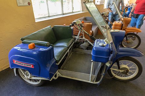 Blue three-wheeler from the side with its windscreen raised, a dark green two-seat bench and a flat floor. Opens a larger view
