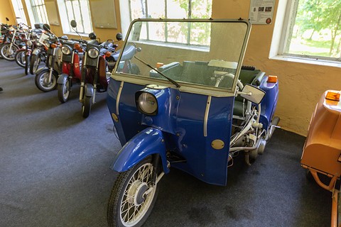 Front of a blue three-wheeler with a single spoked front wheel, a round headlamp and a tall windscreen, scooters in a row behind. Opens a larger view