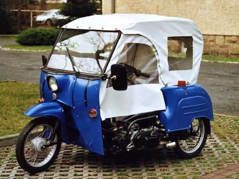 Blue three-wheeler with a white hood and side curtain, a large mirror and an exposed engine, on paved grass. Opens a larger view
