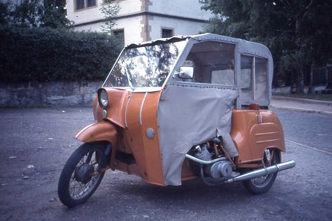 Orange three-wheeled invalid carriage with a grey fabric hood and side curtain, parked in a street at dusk.