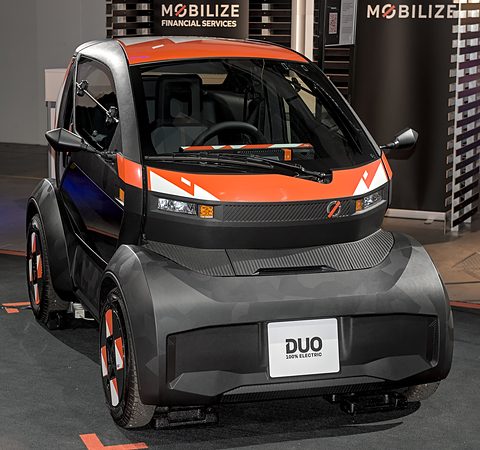 Dark grey tiny two-seater with an orange and white bonnet stripe and a DUO plate, on a show floor. Opens a larger view