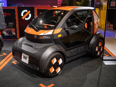 Dark grey tiny two-seater with orange stripes and chequered wheel covers on a dark carpeted stand. Opens a larger view