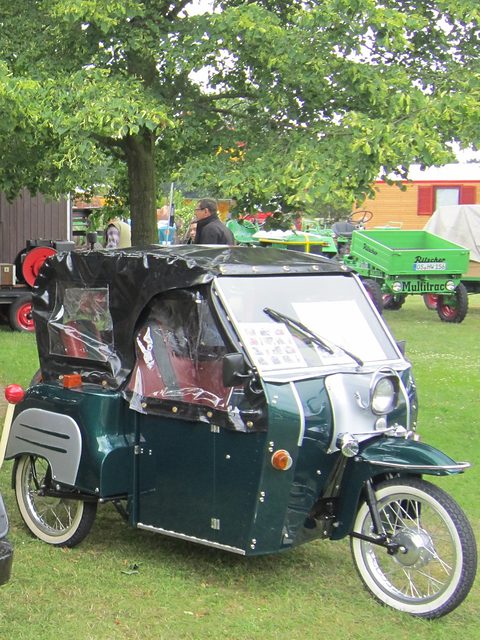 Dark green three-wheeler with a black fabric hood and clear side windows, parked on grass under a tree. Opens a larger view