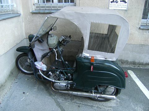 Side view of a dark green three-wheeler with a pale grey fabric hood and clear rear window, against a building wall. Opens a larger view