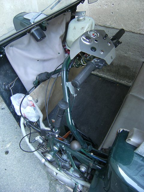 View from above into a dark green three-wheeler: handlebars with control levers, a round speedometer and a ribbed floor. Opens a larger view