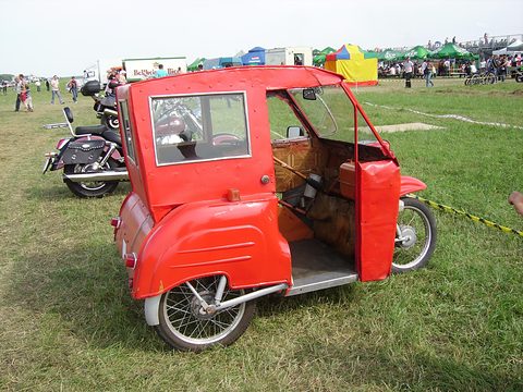 Red three-wheeler from the rear quarter with its side door open to show the seat and floor, a crowd and tents in the distance. Opens a larger view