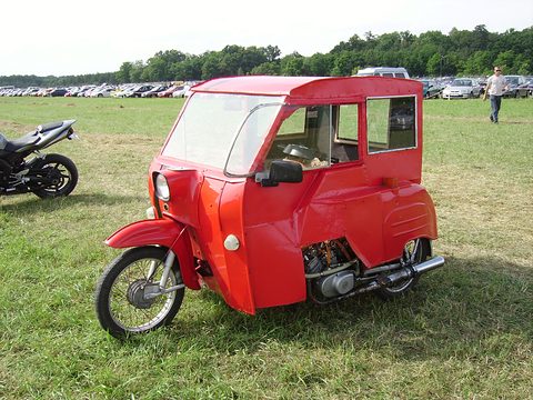 Bright red three-wheeler with a boxy hardtop and a dark stripe, parked on grass with motorcycles and cars behind. Opens a larger view